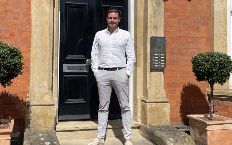 Picture of a white male wearing a white collared shirt and light coloured trousers stood outside the doorway of a red brick building