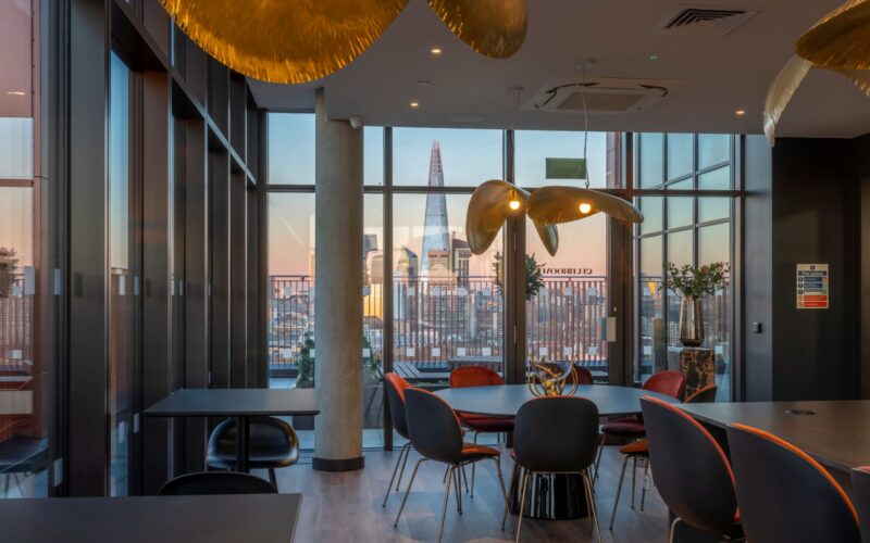 Interior of a building featuring columns and windows, hanging lights, a series of desks and chairs and a view of office towers in the distance