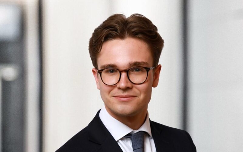A man with dark brown hair wearing glasses and a dark suit with a white shirt and tie
