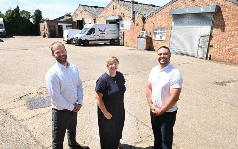 Two men and a woman stood in a yard in front of a warehouse building
