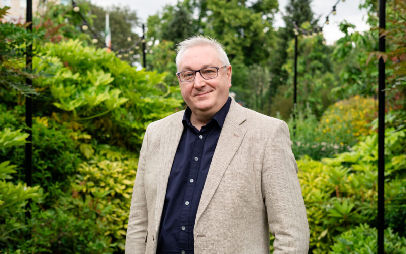 A man wearing spectacles, a light coloured jacket and a dark collared shirt