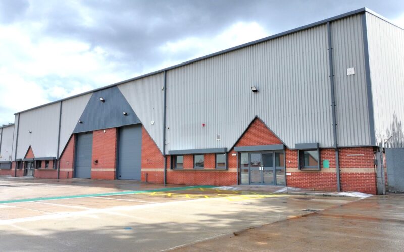 A row of brick and steel cladded industrial buildings with car parking and yard space in the foreground