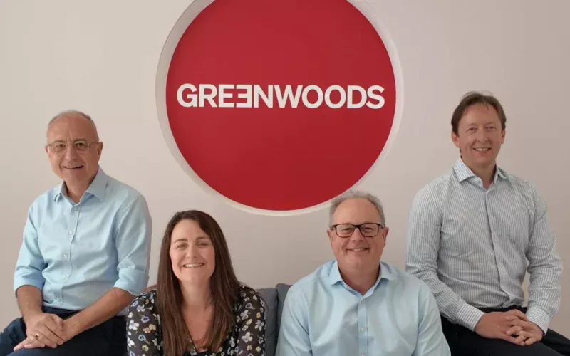 Three men and a women with a large red Greenwoods logo behind them