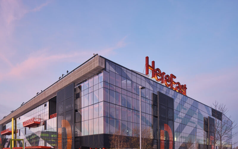 A modern office building featuring lots of external glazing with a large red Here East sign on top