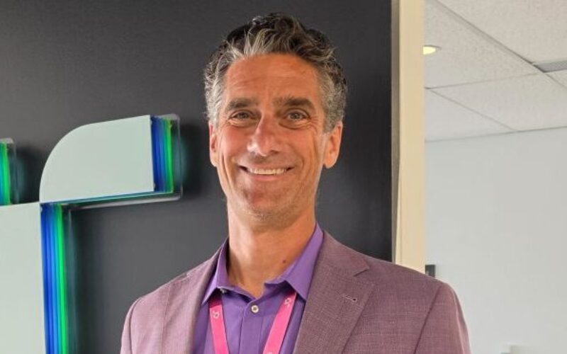 Man wearing a purple shirt and jacket stood next to a company logo