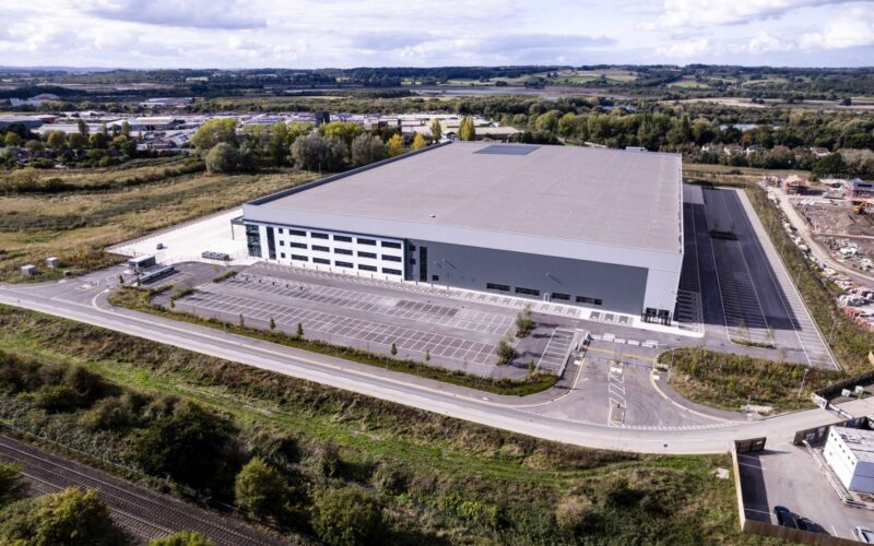 Aerial view of a large modern industrial building surrounded by a wooded area and grassland