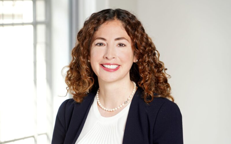A smiling white woman with long curly brown hair and red lipstick wearing a white top, a blue jacket and a pearl necklace