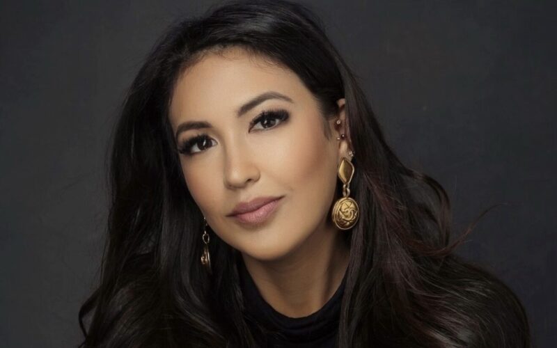 A woman with dark long hair wearing a dark top and gold earrings
