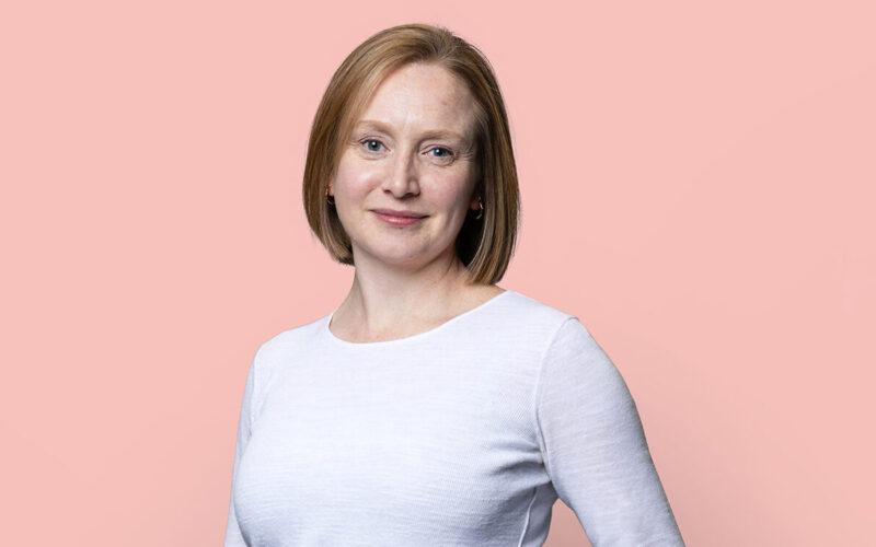 Liz Turner has a bob and is stood against a peach background. She is wearing a light top and dark trousers