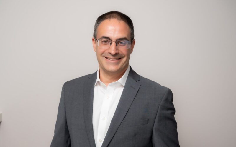 A smiling man with short hair wearing glasses, a grey suit jacket and a white collared shirt