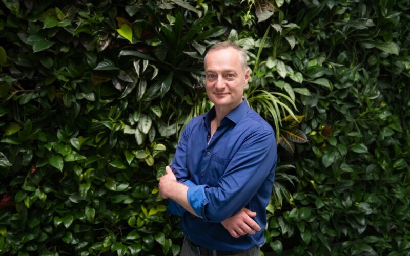 A smiling man wearing a blue shirt stood infront of a green bush