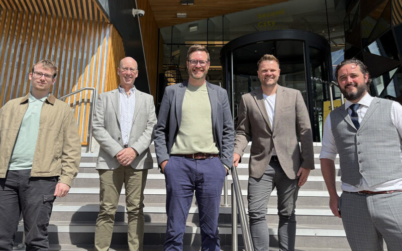 Four smiling men wearing smart clothing stood outside the entrance to a building