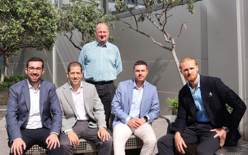 Five men stood outside a building in the sunshine with small trees in the background