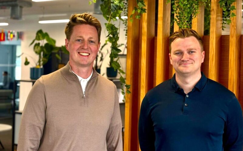 Two white males stood in front of a wooden background with foliage above their heads
