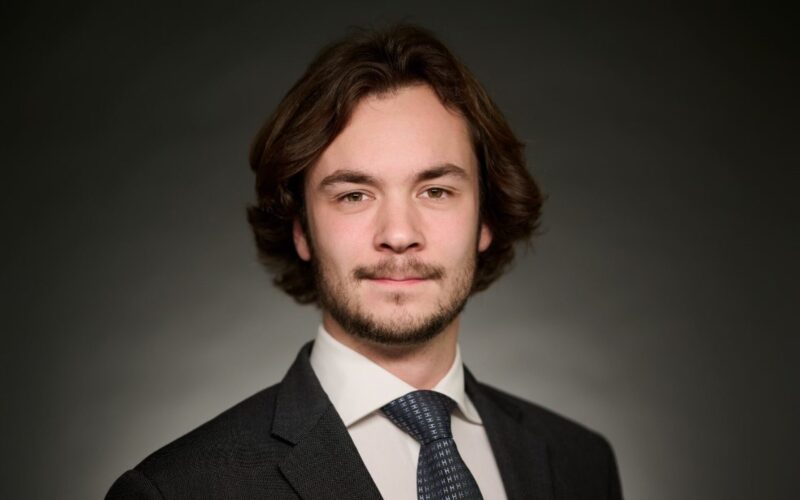 A white male with long brown hair and a beard wearing a dark suit jacket, a white shirt and a dark coloured tie