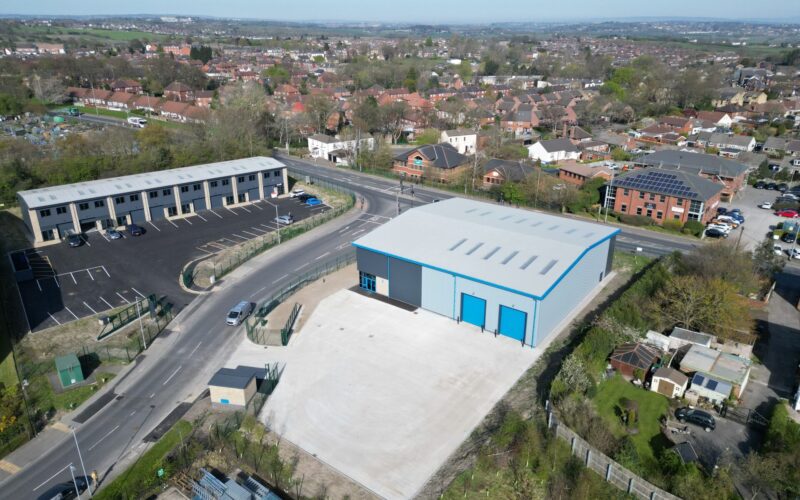 A large industrial unit with blue doors viewed from the air. There is a housing estate behind the site and small office buildings